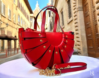 Puede incluir: Un bolso de mano de cuero rojo con un diseño único en forma de abanico y detalles dorados. El bolso tiene una correa de cadena y un asa superior. El bolso está fotografiado con el fondo de una calle de una ciudad italiana.