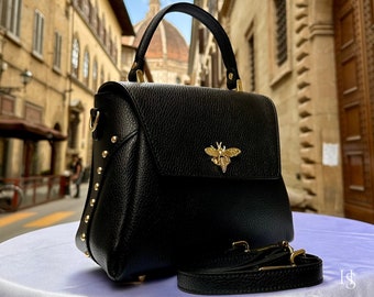 May include: Black leather handbag with a flap closure and a gold bee emblem. The bag has a top handle and a detachable shoulder strap. Gold studs adorn the sides. The text "ITALYLEATHERIT" is visible in the top left corner.