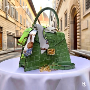 May include: A green crocodile embossed leather handbag with a gold chain strap and a green and white patterned scarf. The bag has a top handle and a gold lock.