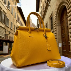 May include: A bright yellow leather handbag with a gold lock and clasp. The bag has a top handle and a detachable shoulder strap. The bag is sitting on a white surface.