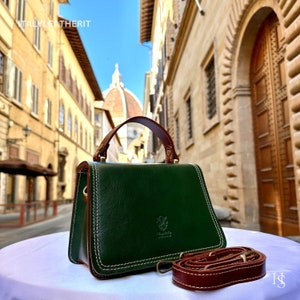 May include: A small, dark green leather handbag with a brown leather handle and strap. The bag has white stitching and a small logo on the front. The bag is sitting on a white surface with a blurred background of a city street.