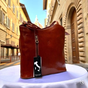 May include: A brown leather tote bag with a zipper and white stitching. The bag has a tag with a map of Italy and the words "GENUINE LEATHER MADE IN ITALY". The bag is on a white surface.