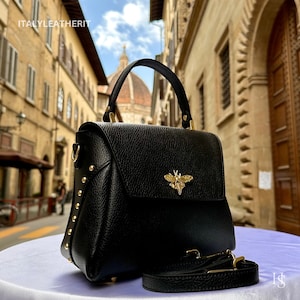 May include: Black leather handbag with a flap closure and a gold bee emblem. The bag has a top handle and a detachable shoulder strap. Gold studs adorn the sides. The text "ITALYLEATHERIT" is visible in the top left corner.