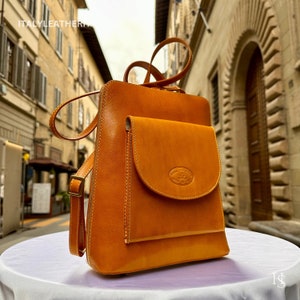 May include: A tan leather backpack with a front pocket and a zipper closure. The bag has a stitched design and a small oval logo on the front pocket.