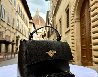 May include: Black leather handbag with a gold-colored bee emblem on the flap. The bag has a top handle and a detachable shoulder strap. The background shows a European street scene.