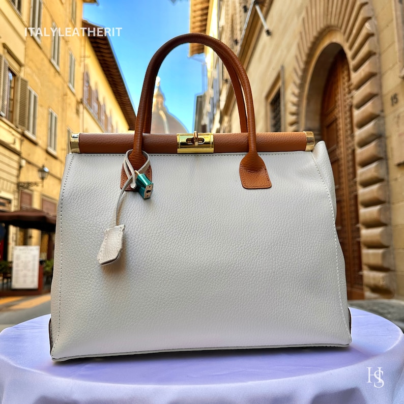 Puede incluir: Un bolso de mano de cuero blanco con ribetes de cuero marr&oacute;n y una cerradura dorada. El bolso est&aacute; sobre una superficie blanca. ITALYLEATHERIT est&aacute; escrito en el edificio del fondo.