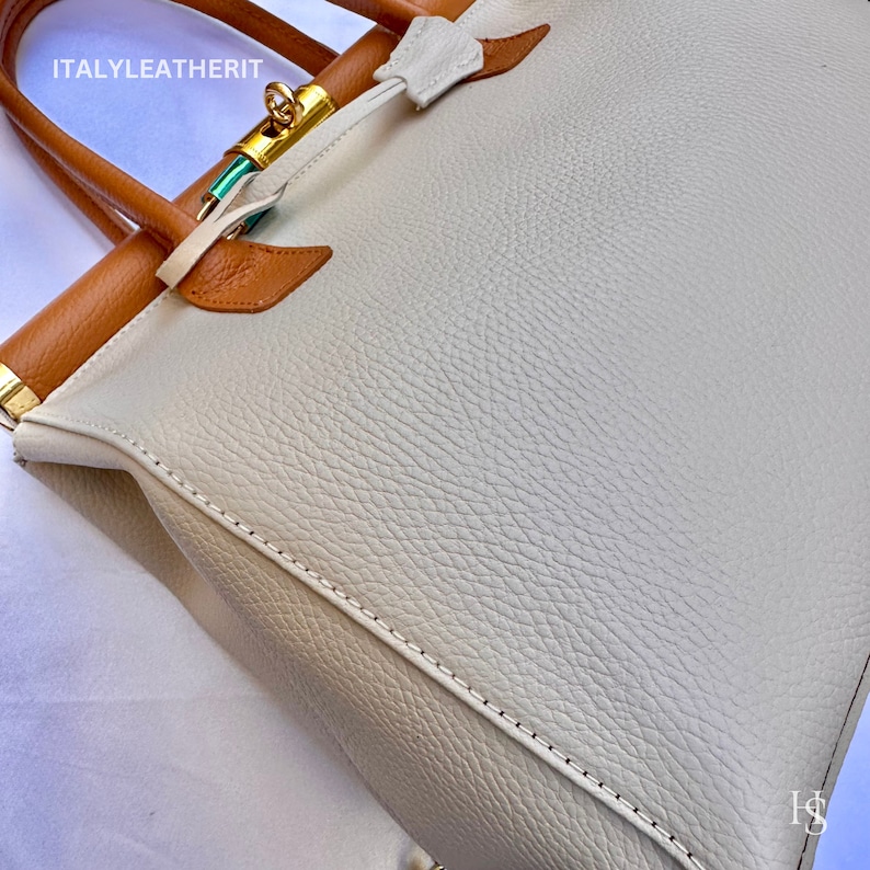 Puede incluir: Un bolso tote de cuero blanco con ribetes de cuero marr&oacute;n y una cerradura dorada. El bolso tiene una superficie texturizada y est&aacute; cosido con hilo blanco.