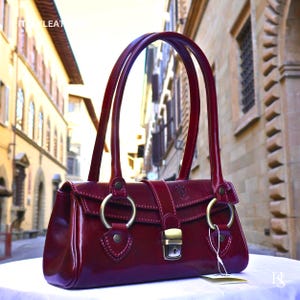 May include: A deep burgundy handbag with a glossy finish, featuring two tall handles, two large circular brass-toned rings at the base of the handles, and a central brass-toned metal clasp closure. The bag has visible stitching and a small tag.