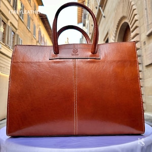 May include: A brown leather tote bag with a top handle and a logo on the front. The bag is sitting on a white surface.