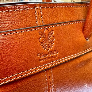 May include: Close-up of a brown leather bag with a stitched design. The bag has a logo that says "Vera Pelle Made in Italy" and a fleur-de-lis symbol.