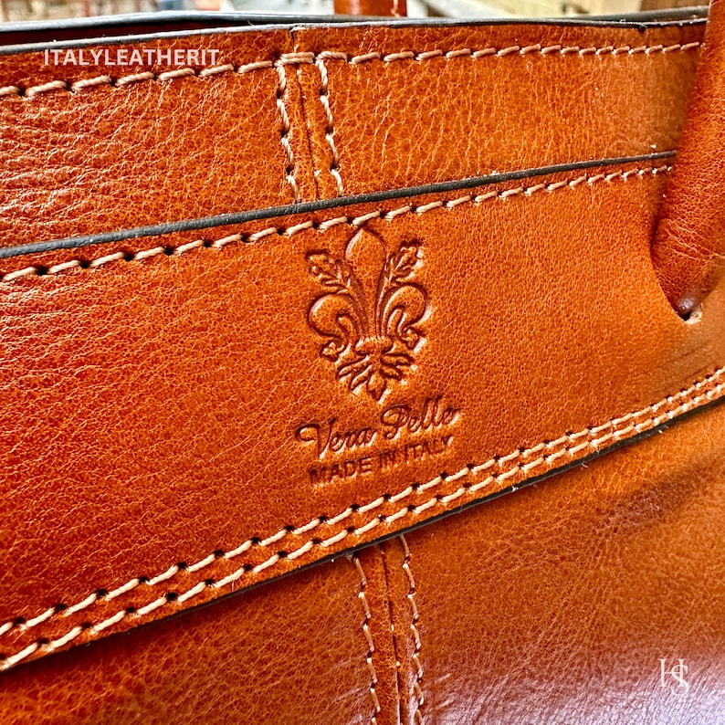 May include: Close-up of a brown leather bag with a stitched design. The bag has a logo that says "Vera Pelle Made in Italy" and a fleur-de-lis symbol.