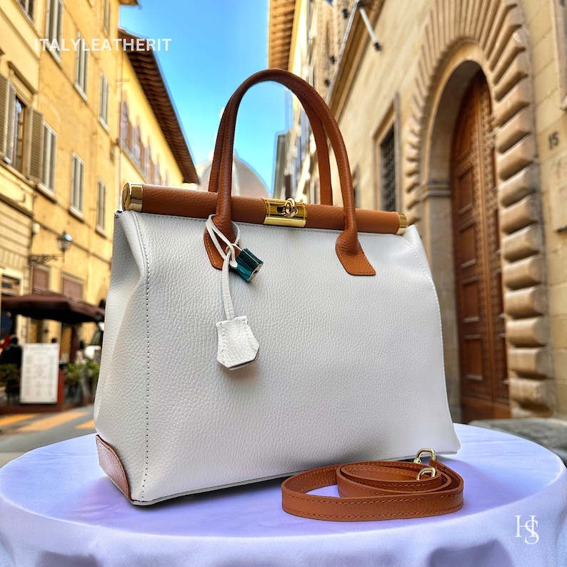 Puede incluir: Un bolso de mano de cuero blanco con ribetes de cuero marr&oacute;n y un cierre dorado. El bolso tiene un asa corta y una correa larga. El bolso est&aacute; sobre una superficie blanca con una correa de cuero marr&oacute;n.