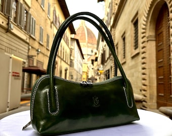 May include: A dark green leather handbag with a curved shape and a zipper closure. The bag has a top handle and a small, embossed logo on the front. The bag is photographed against a backdrop of a city street.