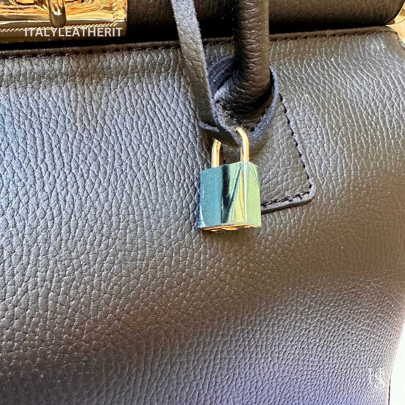 K&ouml;nnte beinhalten: Eine schwarze Lederhandtasche mit einem goldenen Schloss und Schl&uuml;ssel. Die Tasche hat eine strukturierte Oberfl&auml;che und ein kleines Etikett mit der Aufschrift "ITALY LEATHERIT".