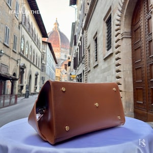Puede incluir: Un bolso de mano de cuero marr&oacute;n con tachuelas doradas. El bolso es rectangular y tiene un dise&ntilde;o estructurado. La imagen incluye el texto "ITALY LEATHER".