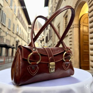 May include: A brown leather handbag with two arched handles and gold-colored hardware. The bag features a rectangular shape with a flap closure and a lock. The stitching is visible in a lighter color, and the bag has a glossy finish. The text "ITALY LEATHER" is visible.