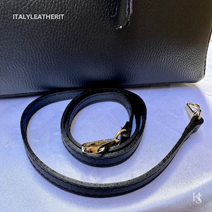 K&ouml;nnte beinhalten: Schwarze Lederschultertasche mit goldfarbenem Metallverschluss und einem schwarzen, verstellbaren Schulterriemen. Der Riemen ist aus dem gleichen Leder wie die Tasche.