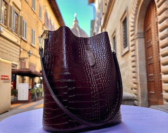 Puede incluir: Un bolso tipo cubo de color burdeos intenso con un acabado texturizado de estampado de cocodrilo. El bolso presenta una forma cilíndrica estructurada, una correa de hombro única con herrajes metálicos dorados y el texto ITALYLEATHERIT arriba.