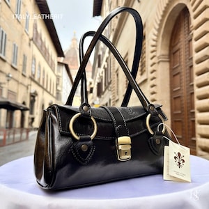 May include: A black leather handbag with gold-tone hardware, including large circular rings and a clasp closure. The bag has two arched handles and a small tag with a floral emblem. The background shows a European street scene.