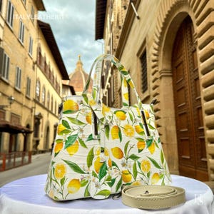 May include: White handbag featuring a lemon and leaf design. The bag has a top handle and a detachable shoulder strap. The background shows a European street scene. The text "ITALYLEATHERIT" is visible on the top left.