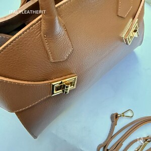 Puede incluir: Un bolso de mano de cuero marr&oacute;n con herrajes dorados. El bolso tiene un asa superior, una correa para el hombro y un cierre rectangular. La textura del cuero es visible. El texto "ITALYLEATHERIT" est&aacute; impreso en el bolso.