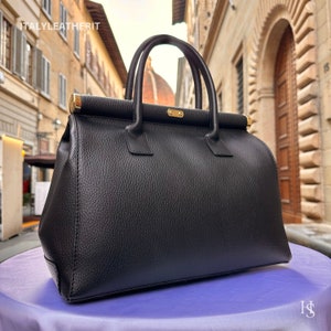 K&ouml;nnte beinhalten: Eine schwarze Ledertasche mit goldenen Beschl&auml;gen. Die Tasche hat einen oberen Henkel und eine strukturierte Form.