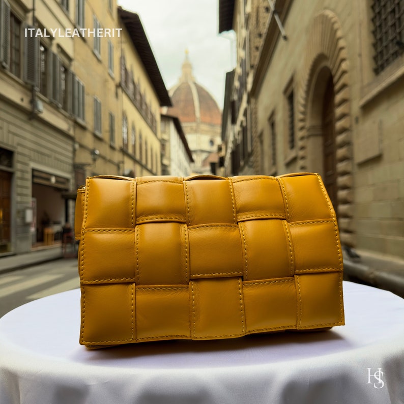 Puede incluir: Un bolso de mano de cuero tejido de color amarillo mostaza. El bolso es rectangular con un dise&ntilde;o acolchado de tejido de cesta. Est&aacute; colocado sobre una superficie blanca, con una escena de calle borrosa en el fondo. El texto "ITALYLEATHERIT" es visible.