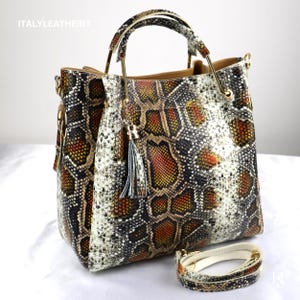 May include: A handbag featuring a snakeskin pattern in shades of orange, black, and white. The bag has gold-tone metal handles, a hanging tassel charm, and a detachable shoulder strap. The text ITALYLEATHERIT is visible in the top left corner.