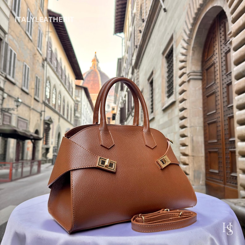 Puede incluir: Un bolso de mano de cuero marr&oacute;n con asa superior y correa de hombro desmontable. El bolso presenta herrajes dorados y un dise&ntilde;o estructurado. El texto "ITALYLEATHER" es visible en la esquina superior izquierda.