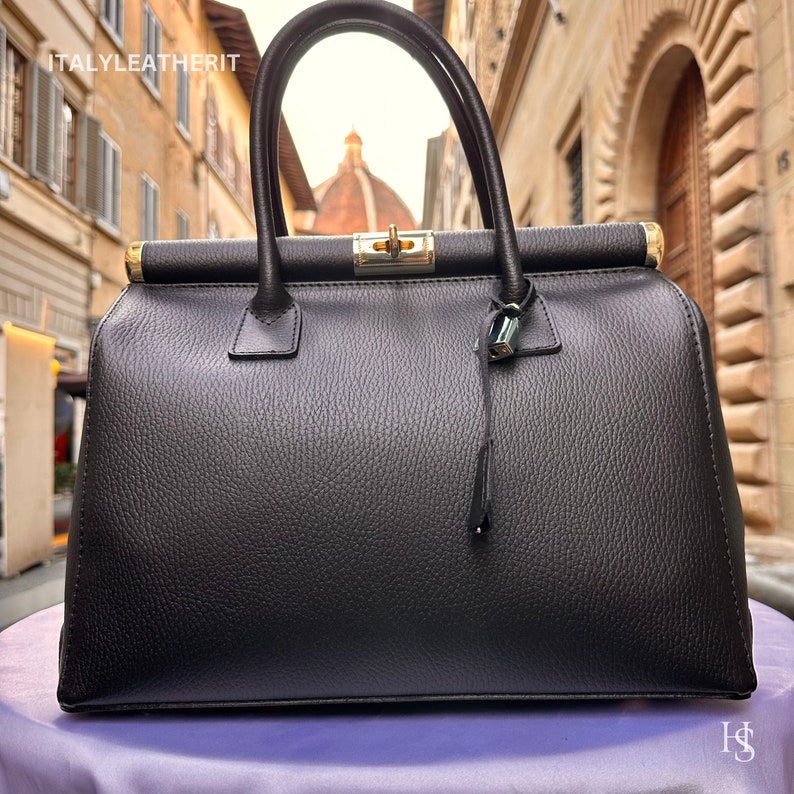 K&ouml;nnte beinhalten: Eine schwarze Ledertasche im Arzt-Stil mit goldenen Beschl&auml;gen und einem kleinen Schloss. Die Tasche hat eine strukturierte Lederoberfl&auml;che und ist geschlossen.