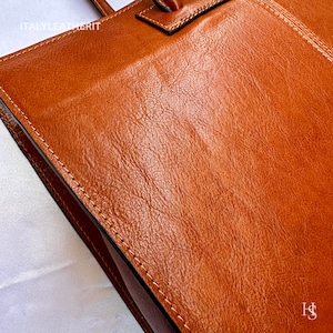 May include: Close-up of a brown leather tote bag with the text "ITALY LEATHERIT" on the front. The bag has a stitched seam around the edge.