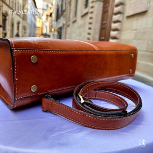 May include: A brown leather handbag with a matching shoulder strap. The bag has a flap closure and a stitched design. The strap is folded and placed next to the bag.