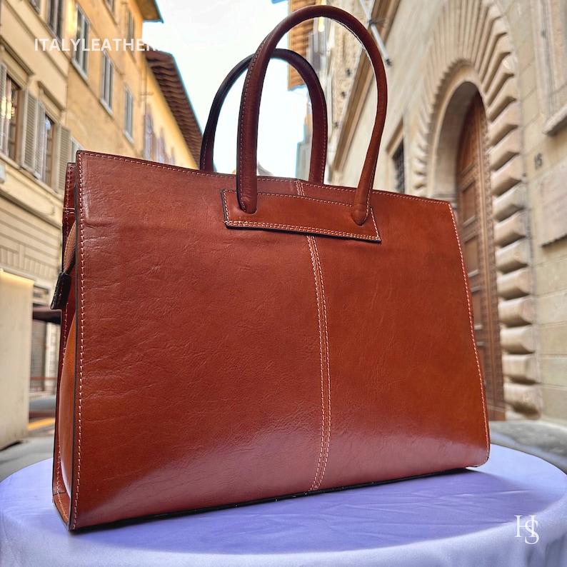 May include: A brown leather tote bag with a top handle and a zip closure. The bag features a stitched design and is crafted from high-quality leather.