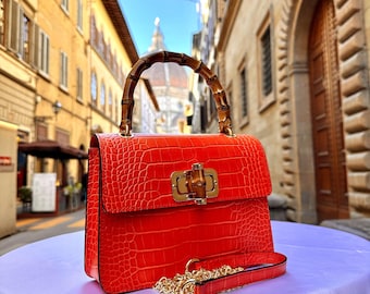 May include: A bright red handbag with a crocodile-effect texture. It has a bamboo handle and a gold-coloured clasp. A matching red strap with a gold chain is also included. The bag is set against a blurred city backdrop.