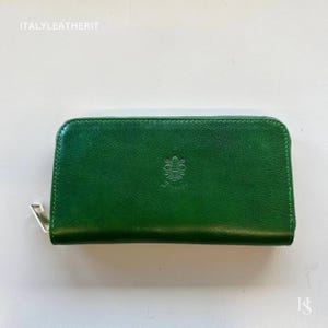 Pode incluir: Uma carteira de couro verde com zíper prateado. A carteira tem um emblema floral em relevo e o texto "ITALYLEATHERIT". A carteira retangular tem uma parte superior arredondada.