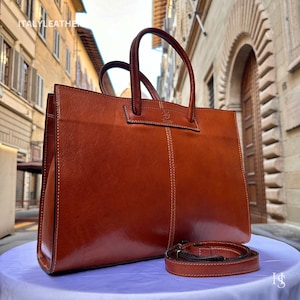 May include: A brown leather tote bag with a top handle and a detachable shoulder strap. The bag has a simple design with a single flap and a stitched seam down the centre.