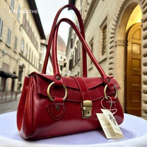 May include: A vibrant red leather handbag with two top handles and gold-tone metal hardware. The bag has a flap closure with a buckle and a keyhole lock. A tag is attached to the bag with text.