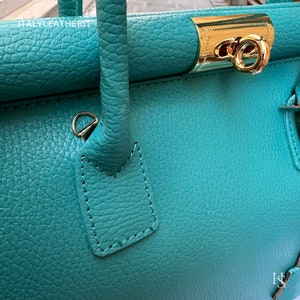 May include: Close-up of a teal leather handbag with a gold clasp and a gold ring handle. The bag has a textured surface and is stitched with a dark thread.