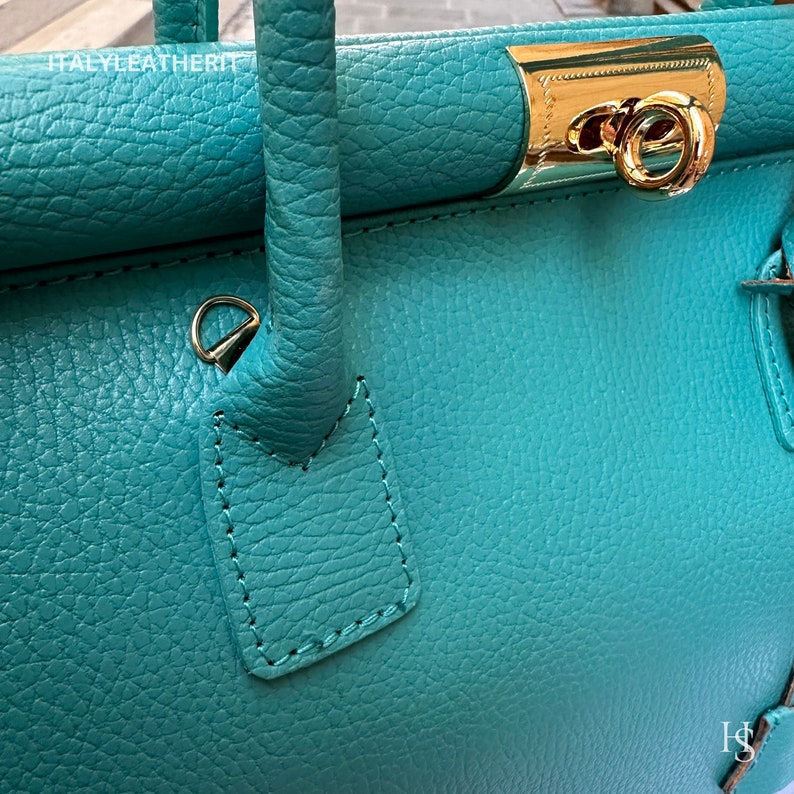 May include: Close-up of a teal leather handbag with a gold clasp and a gold ring handle. The bag has a textured surface and is stitched with a dark thread.