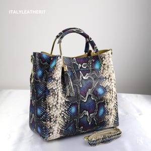 May include: A handbag featuring a snakeskin pattern in shades of blue, purple, black, and off-white. The bag has two gold-tone metal handles, a matching detachable shoulder strap, and a small tassel accent. The text ITALYLEATHERIT is visible.