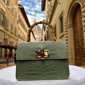 May include: A green crocodile embossed leather handbag with a bamboo handle and gold hardware. The bag has a flap closure with a gold buckle and a top handle.