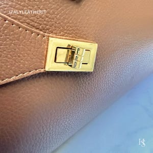 Puede incluir: Primer plano de un bolso de mano de cuero marr&oacute;n con un cierre rectangular dorado. El cierre est&aacute; centrado y tiene un mecanismo de cierre a presi&oacute;n. El cuero tiene un aspecto texturizado. El texto "ITALYLEATHERIT" es visible.