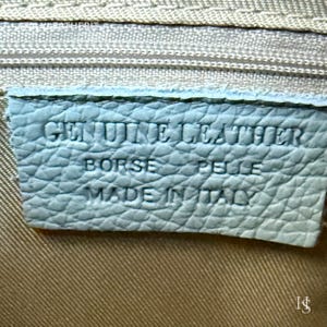 Puede incluir: Primer plano de una etiqueta de cuero azul claro con texto en relieve. La etiqueta dice "GENUINE LEATHER BORSE PELLE MADE IN ITALY". La etiqueta es rectangular y tiene una superficie texturizada. El fondo es de tela beige claro.