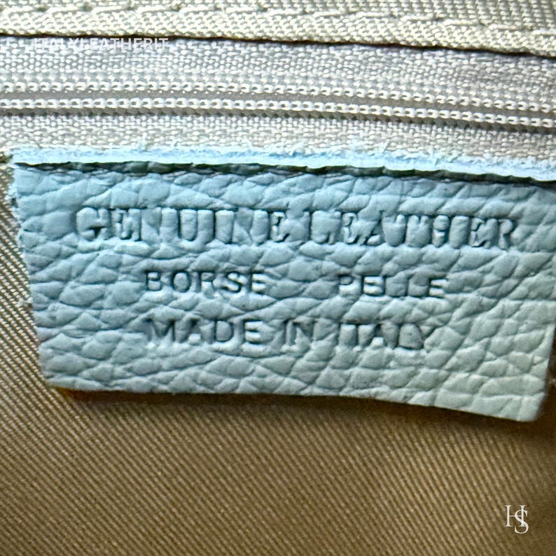 Puede incluir: Primer plano de una etiqueta de cuero azul claro con texto en relieve. La etiqueta dice "GENUINE LEATHER BORSE PELLE MADE IN ITALY". La etiqueta es rectangular y tiene una superficie texturizada. El fondo es de tela beige claro.