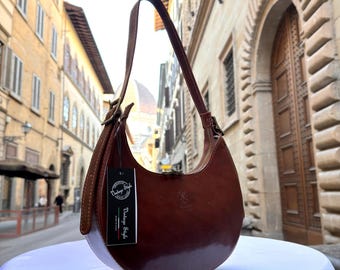 Könnte beinhalten: Eine braune Leder-Umhängetasche mit geschwungener Form. Die Tasche hat einen langen Riemen und einen Reißverschluss. Ein Etikett lautet "Vintage Style, Made in Italy". Die Tasche wird auf einer weißen Oberfläche mit einer verschwommenen Straße im Hintergrund präsentiert.