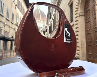 Könnte beinhalten: Eine halbmondförmige, braune Lederhandtasche mit passendem Schulterriemen. Die Tasche hat einen Reißverschluss und einen Griff. Ein Etikett mit der italienischen Flagge ist angebracht. Die Tasche wird auf einer weißen Oberfläche präsentiert.