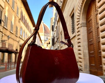 Könnte beinhalten: Eine glänzende, weinrote Leder-Umhängetasche mit geschwungener Form und langem Riemen. Die Tasche hat ein dezentes geprägtes Design und ist vor einem verschwommenen Stadtbildhintergrund platziert. Ein klassisches Accessoire.
