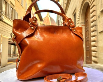 May include: A glossy, orange-brown leather handbag with a top handle and a detachable shoulder strap. The bag features gold-colored hardware and a structured design. The text "ITALY LEATHERIT" is visible in the upper left corner.