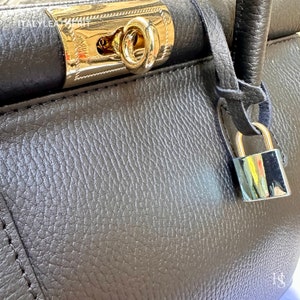 K&ouml;nnte beinhalten: Eine dunkelgraue Ledertasche mit einem goldenen Schloss und einem Schl&uuml;sselloch. Die Tasche hat eine strukturierte Oberfl&auml;che und einen langen Riemen.