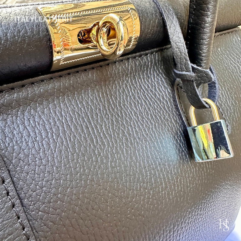 K&ouml;nnte beinhalten: Eine dunkelgraue Ledertasche mit einem goldenen Schloss und einem Schl&uuml;sselloch. Die Tasche hat eine strukturierte Oberfl&auml;che und einen langen Riemen.